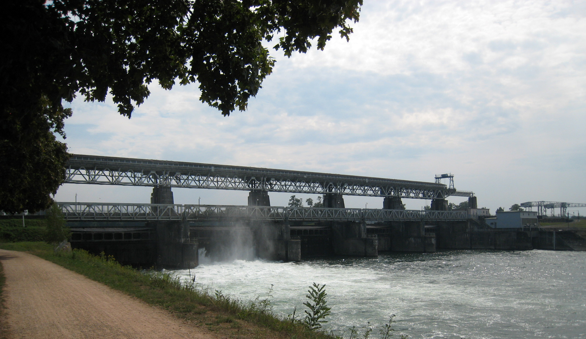 A concrete barrage spanning the Rhine, seen from a gravel path along the bank. Water pours white through the gates below a steel gantry that carries the full width of the river; the powerhouse and a crane stand at the far end.